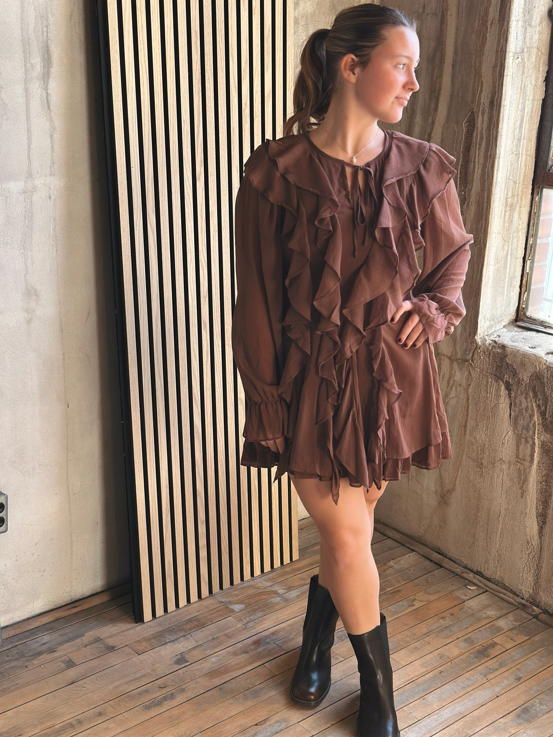 Woman wearing a brown dress with ruffles standing in a room with wooden floor and concrete wall.
