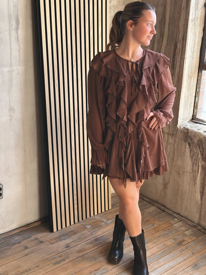 Woman wearing a brown dress with ruffles standing in a room with wooden floor and concrete wall.