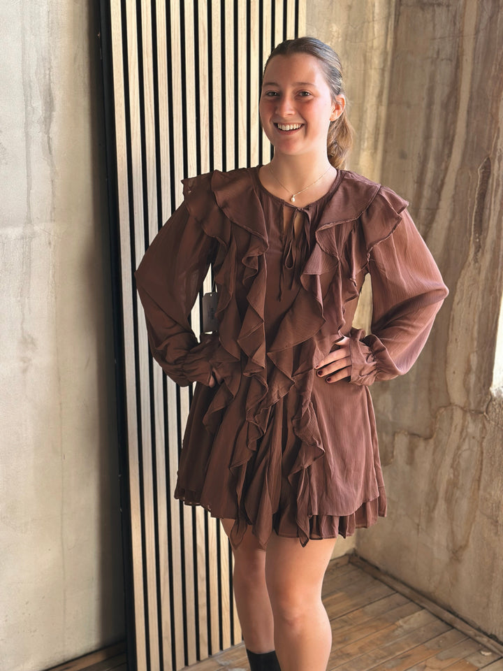 Woman wearing a brown ruffled dress standing against a textured wall.