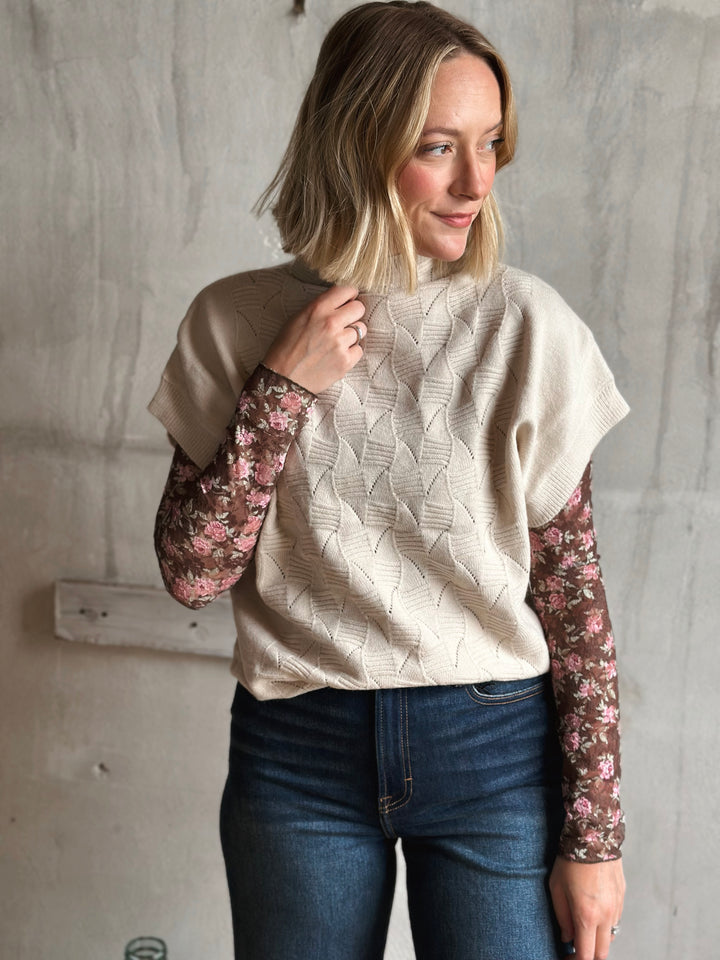 Woman wearing a beige sweater with floral sleeves against a gray wall.