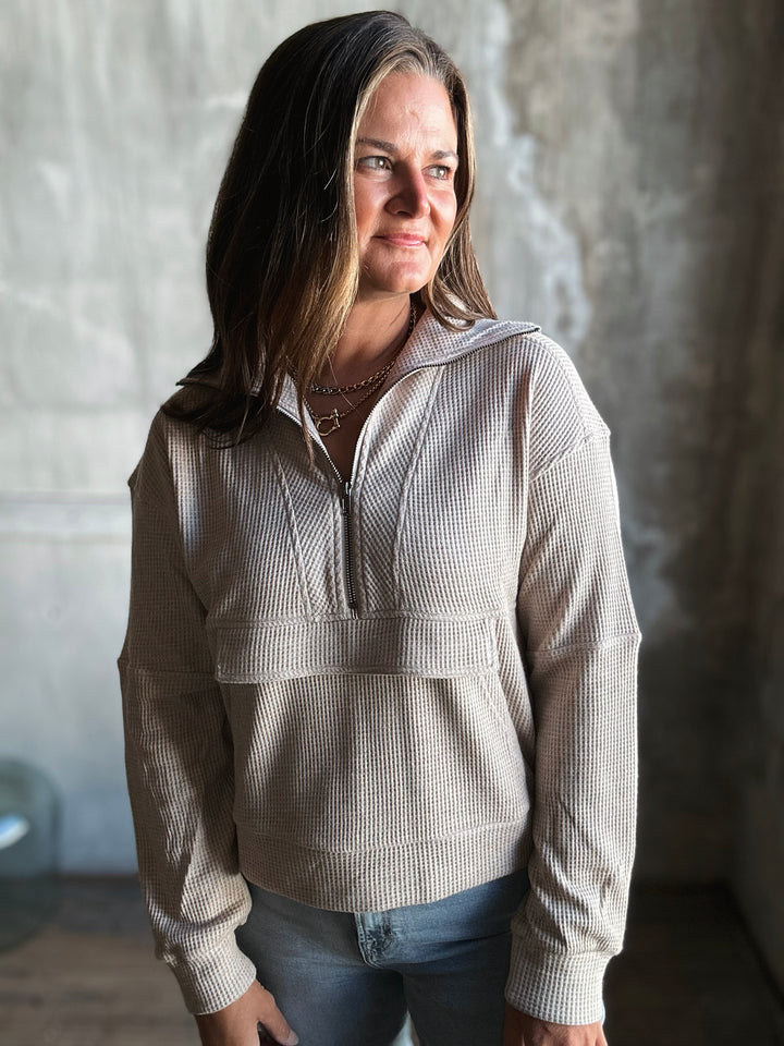 Woman wearing a beige checkered pullover with a hood against a textured wall.