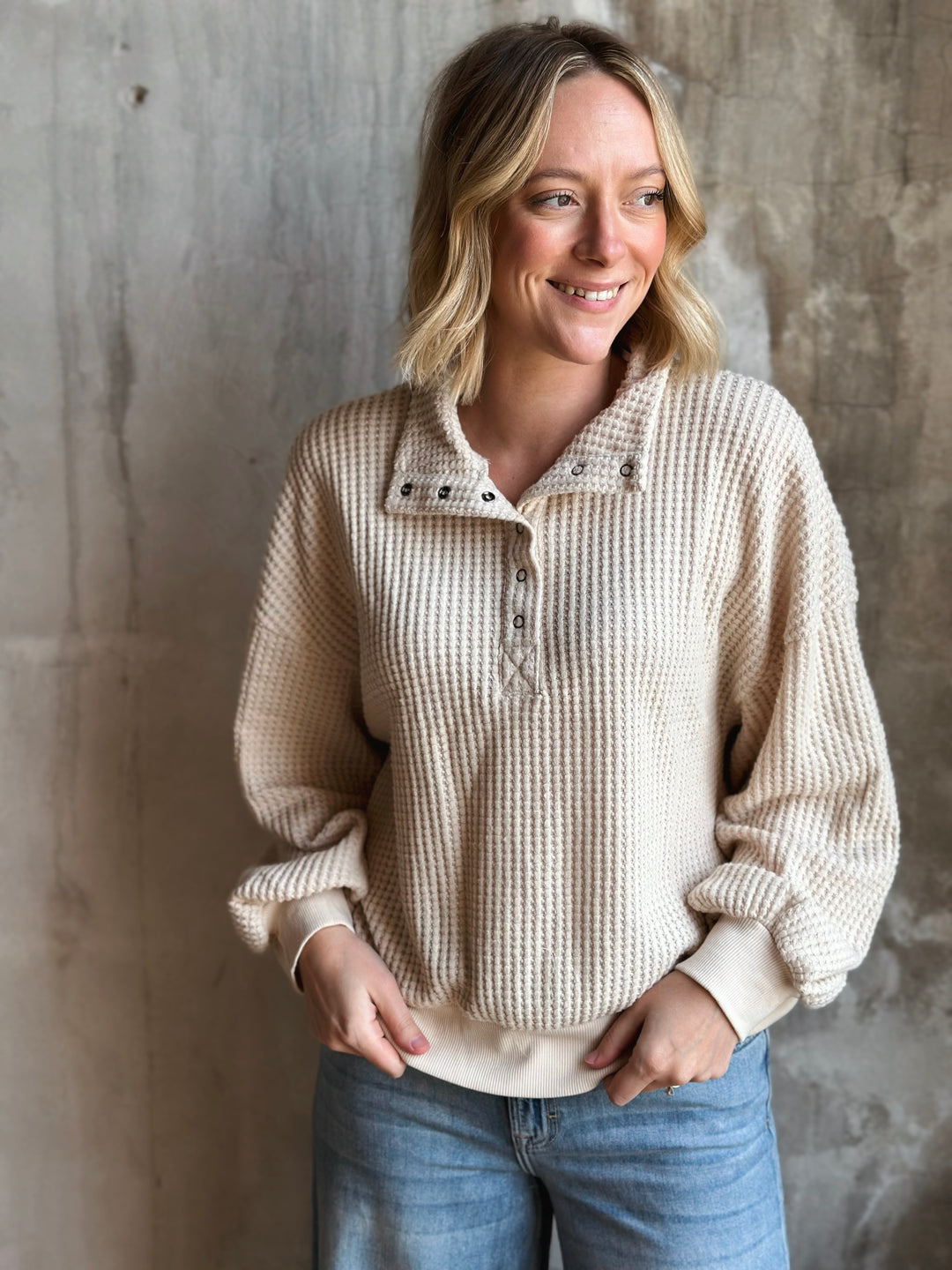 Woman wearing a beige knitted sweater and blue jeans against a textured wall.
