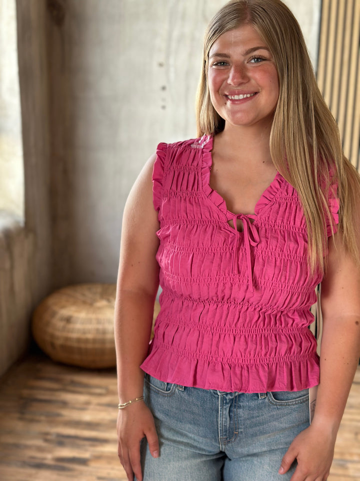  A woman wearing a pink ruffle sleeveless top and blue jeans indoors.
