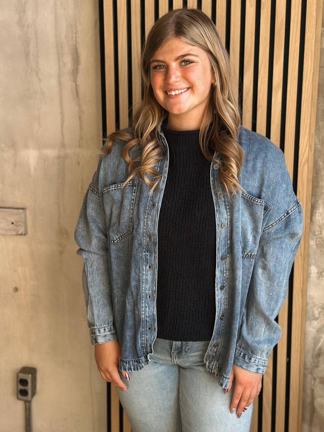Woman wearing a denim jacket over a black top and jeans, standing against a wooden paneled wall.