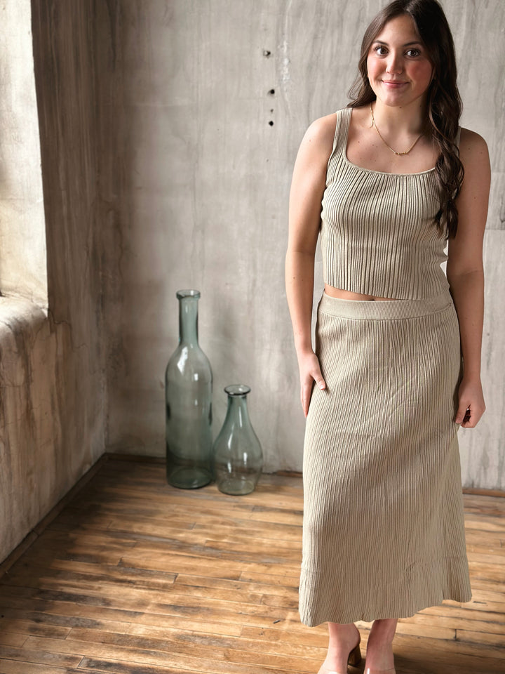 Woman wearing a beige ribbed top and skirt standing in a room with wooden floor and textured walls.