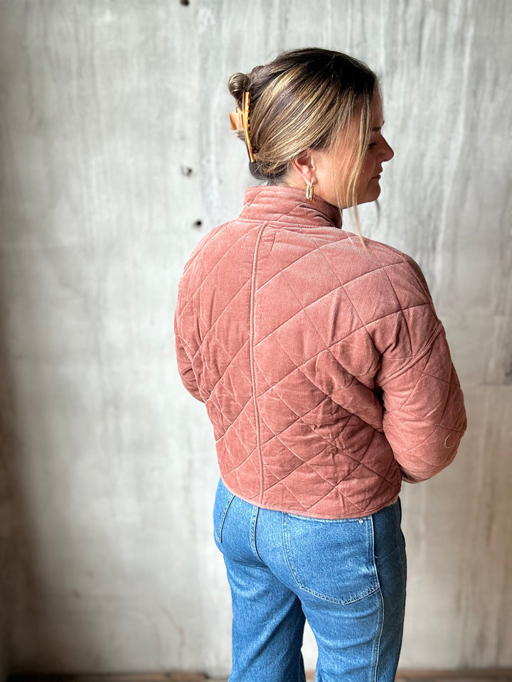 A woman shown from the back wearing a dusty rose quilted corduroy jacket and blue jeans, standing against a textured concrete wall.