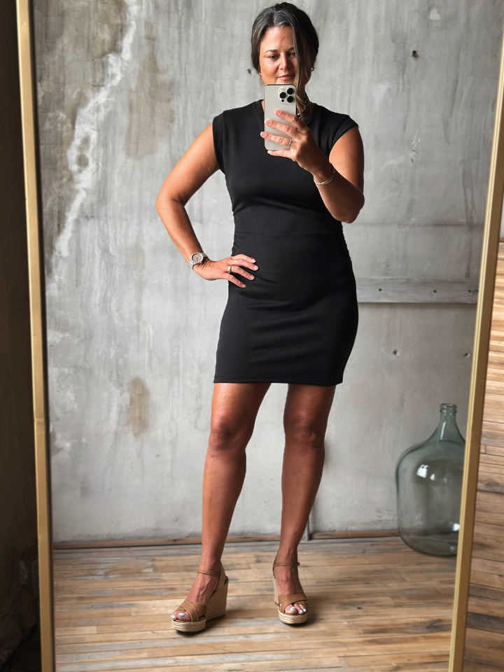 Woman taking a mirror selfie wearing a black dress in a room with wooden floor and concrete wall.