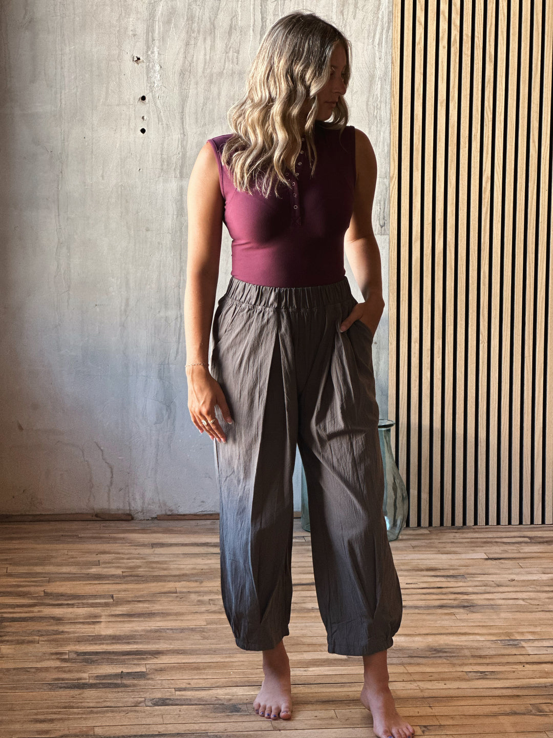 Woman wearing a burgundy top and gray pants standing in a room with wooden flooring and a textured wall.