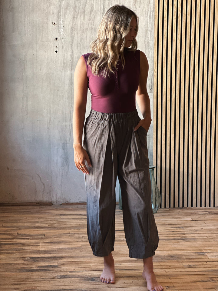 Woman wearing a burgundy top and gray pants standing in a room with wooden flooring and a textured wall.