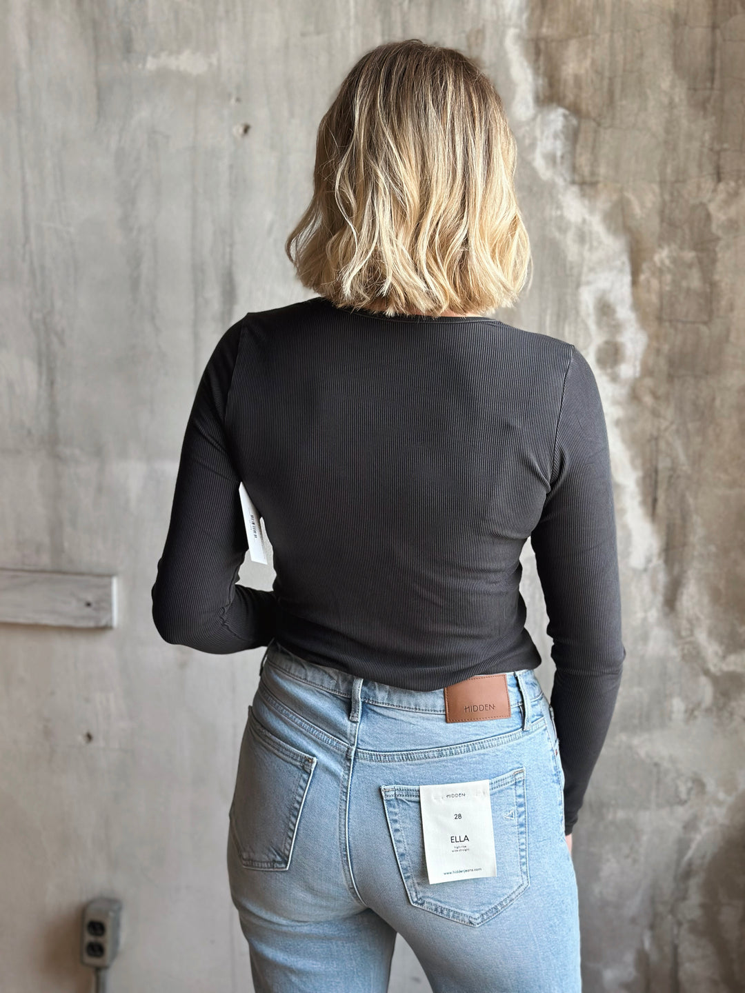 Person wearing a black long-sleeve top and light blue jeans against a gray concrete wall.