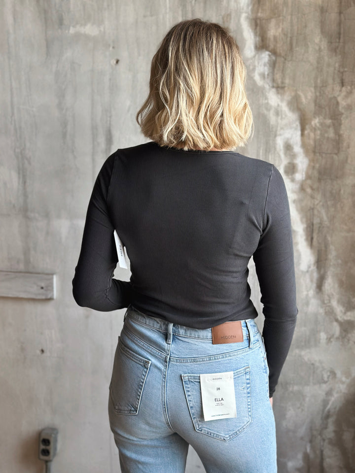 Person wearing a black long-sleeve top and light blue jeans against a gray concrete wall.