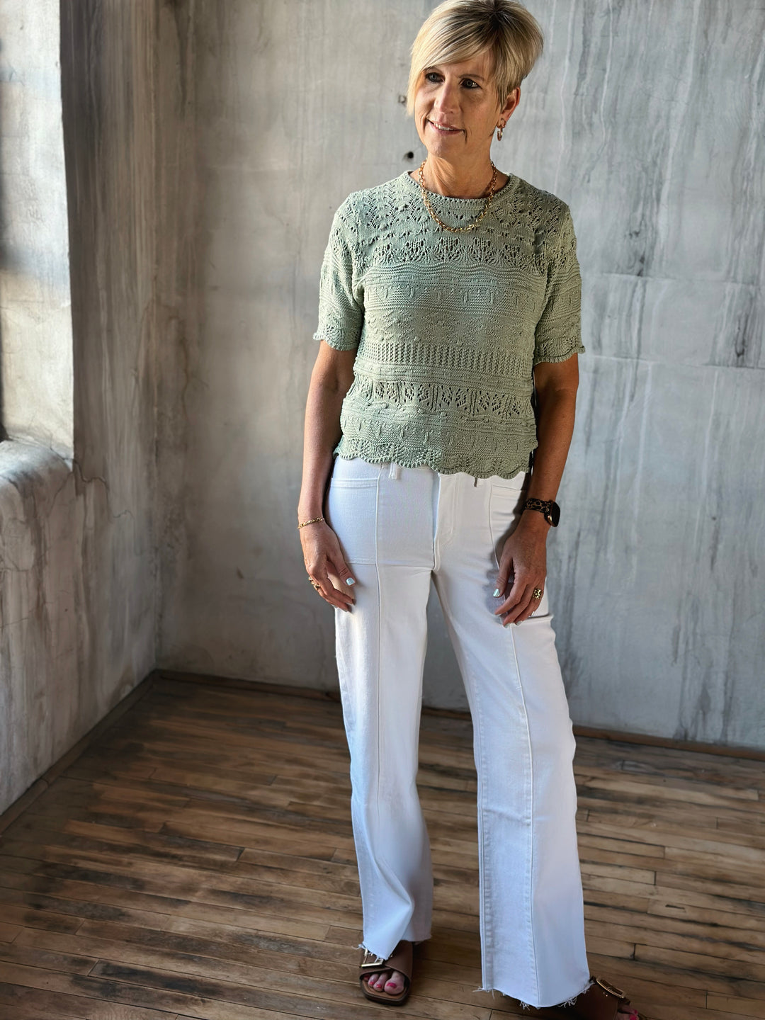 Woman wearing a green lace top and white pants standing in a room with wooden floor and stone walls.