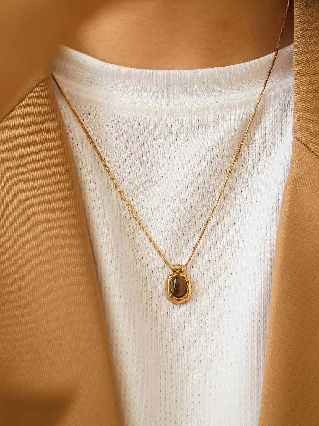 Gold necklace with a pendant worn by a person in a white top and brown jacket.