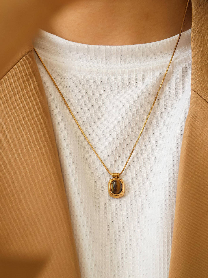 Gold necklace with a pendant worn by a person in a white top and brown jacket.