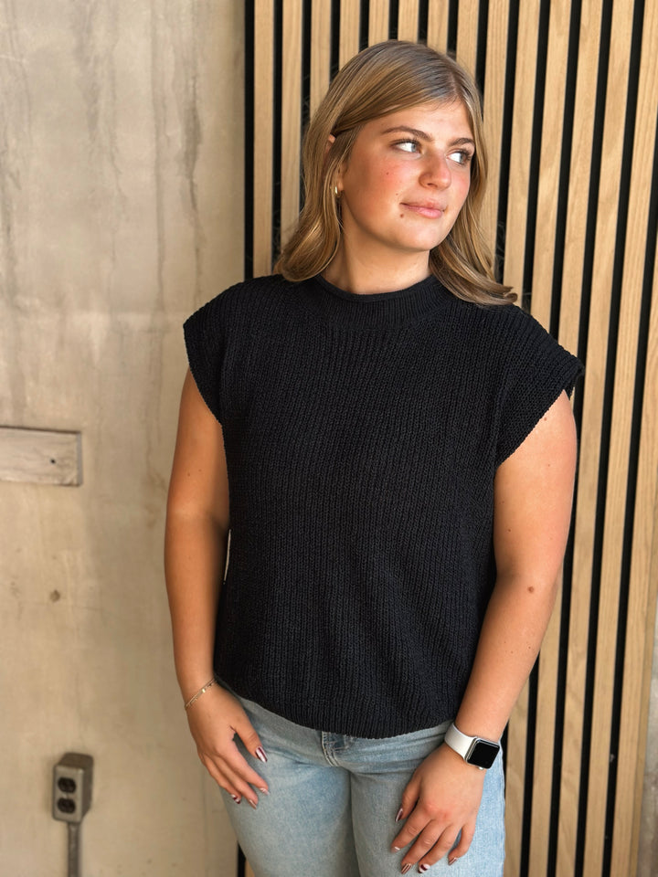 Woman wearing a black sleeveless top and light blue jeans standing against a wooden wall.
