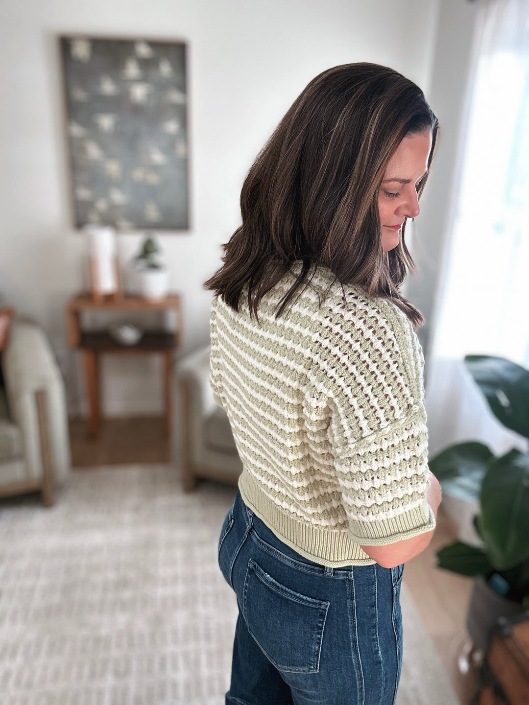 Woman wearing a light green knitted sweater and blue jeans in a living room.