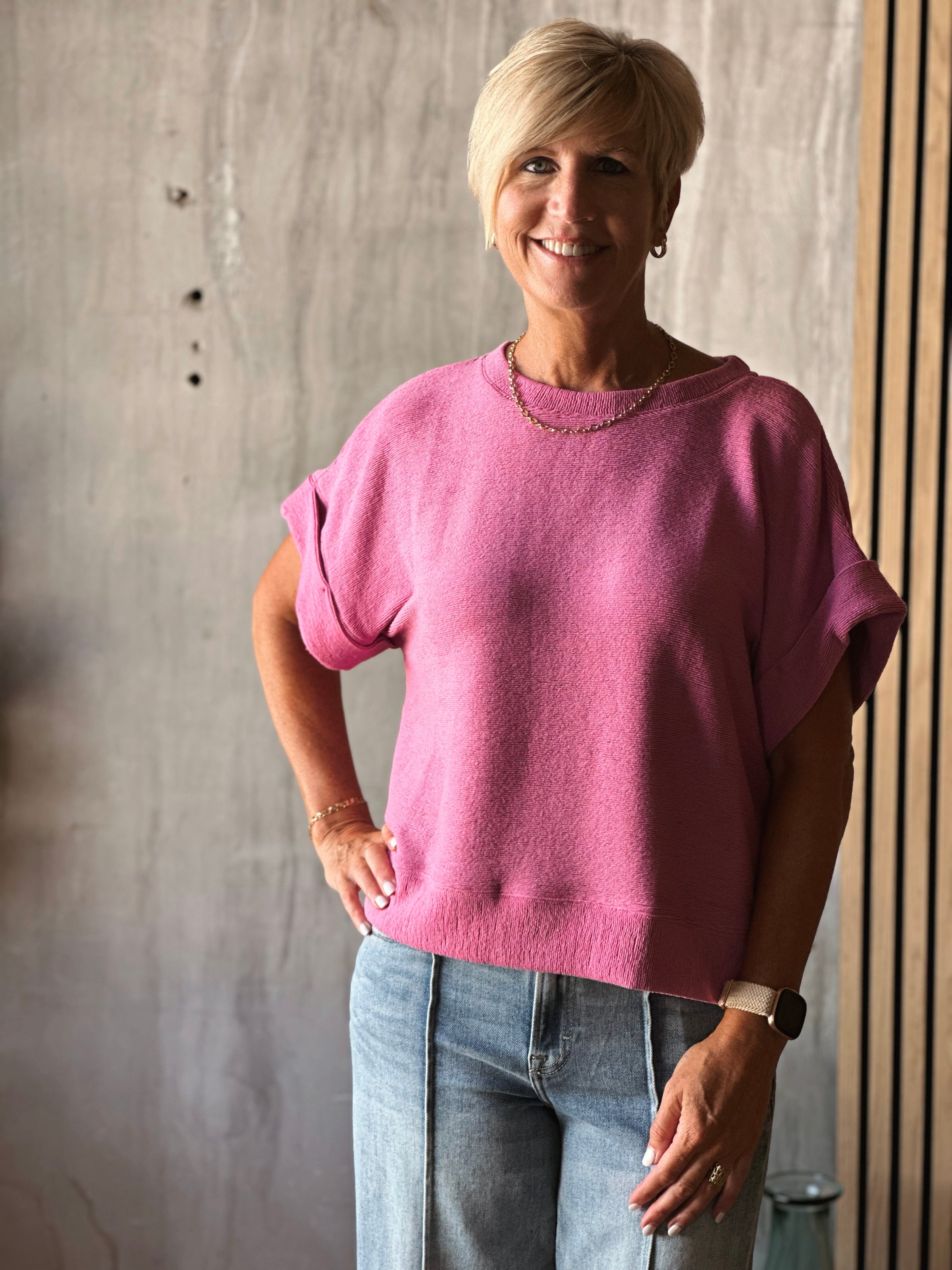 Woman wearing a pink sweater and blue jeans standing against a concrete wall.