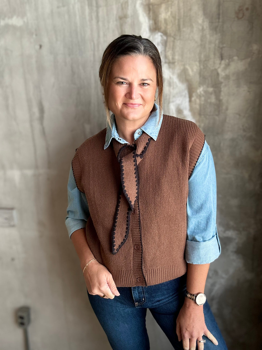 A woman wearing a brown ribbed knit vest with a tie-neck detail over a light blue button-up shirt and dark blue jeans, standing against a textured concrete wall.
