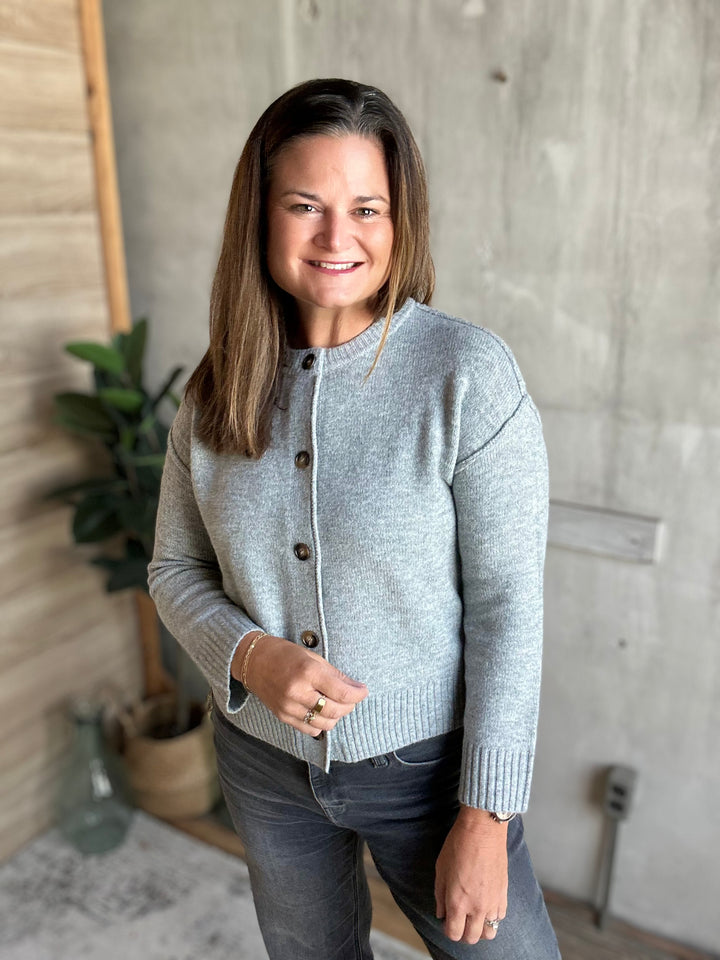 A woman wearing a light gray knit cardigan with dark buttons and dark gray jeans, standing in a modern room with a concrete wall and wood paneling.