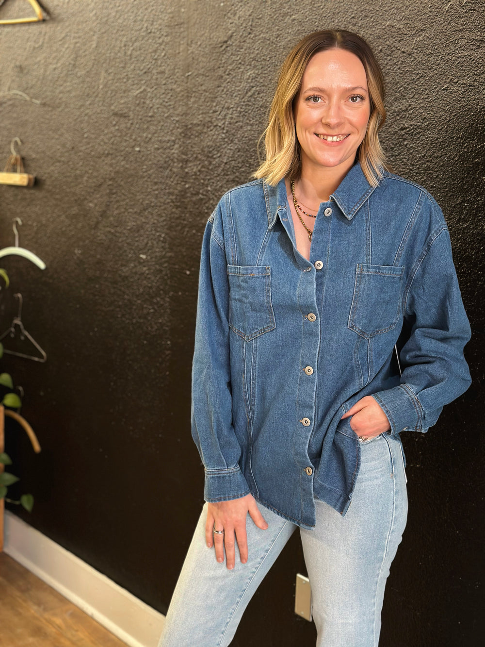 Woman wearing a denim shirt and jeans standing against a dark wall.