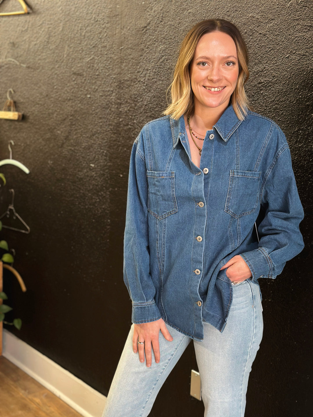 Woman wearing a denim shirt and jeans standing against a dark wall.