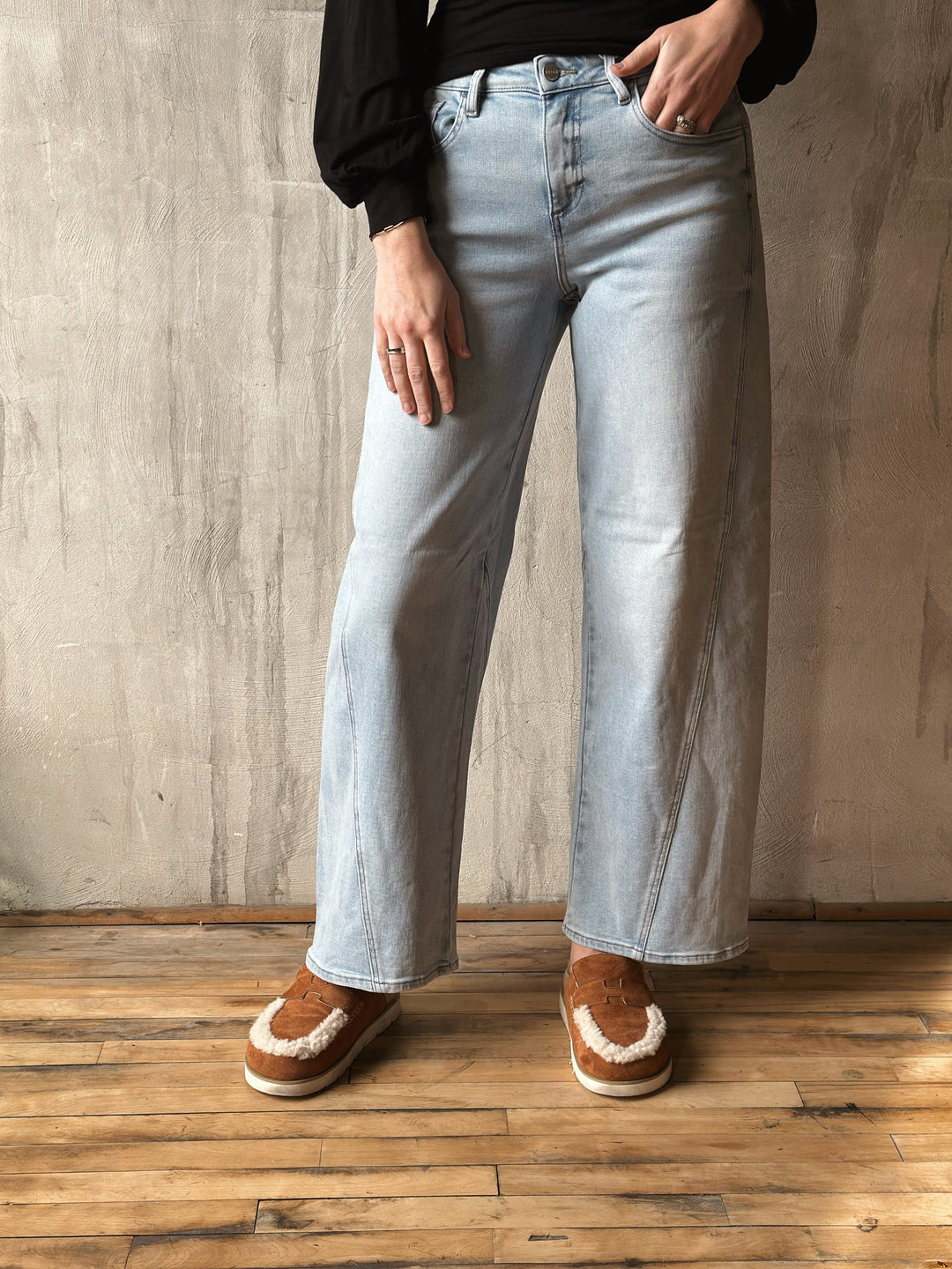 Person wearing light blue jeans and brown shoes against a wooden floor and gray wall background