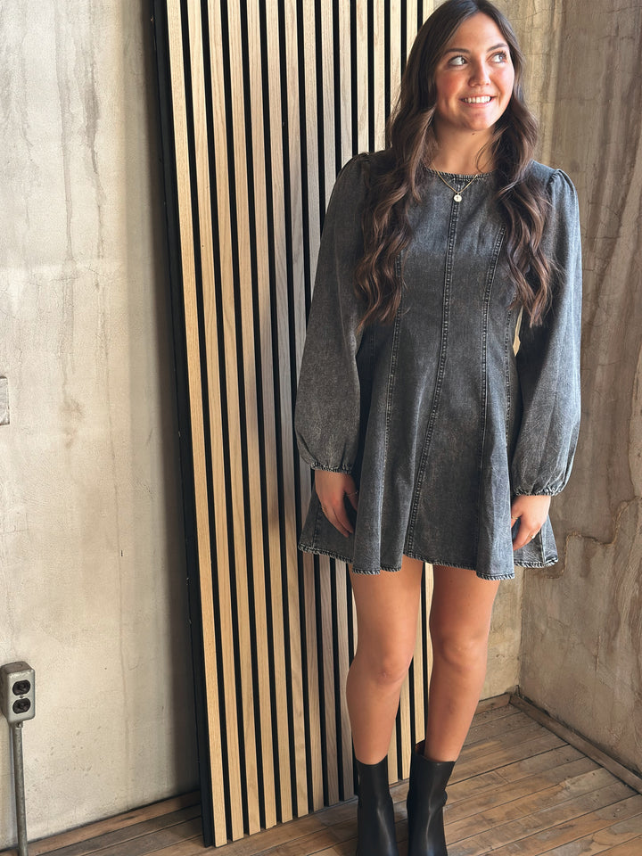 Woman wearing a denim dress standing against a wooden panel wall.