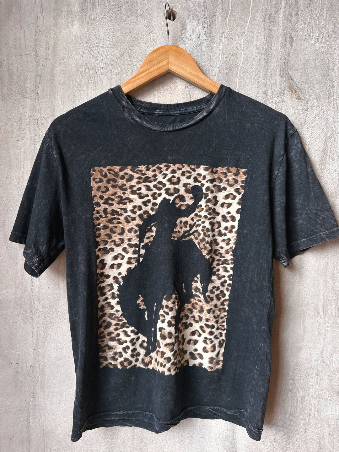 Black t-shirt with a leopard print and horse silhouette on a wooden hanger against a gray concrete wall.