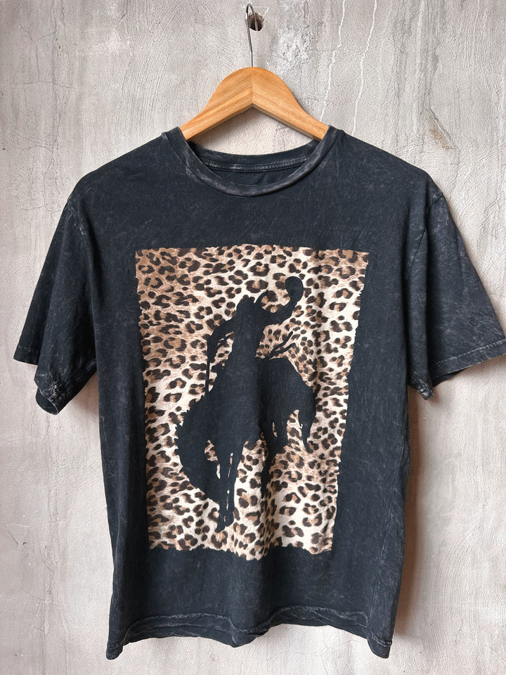 Black t-shirt with a leopard print and horse silhouette on a wooden hanger against a gray concrete wall.