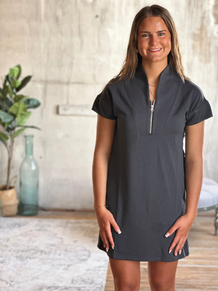 Woman wearing a dark gray dress with short sleeves and a zipper in a room with a plant and vase.