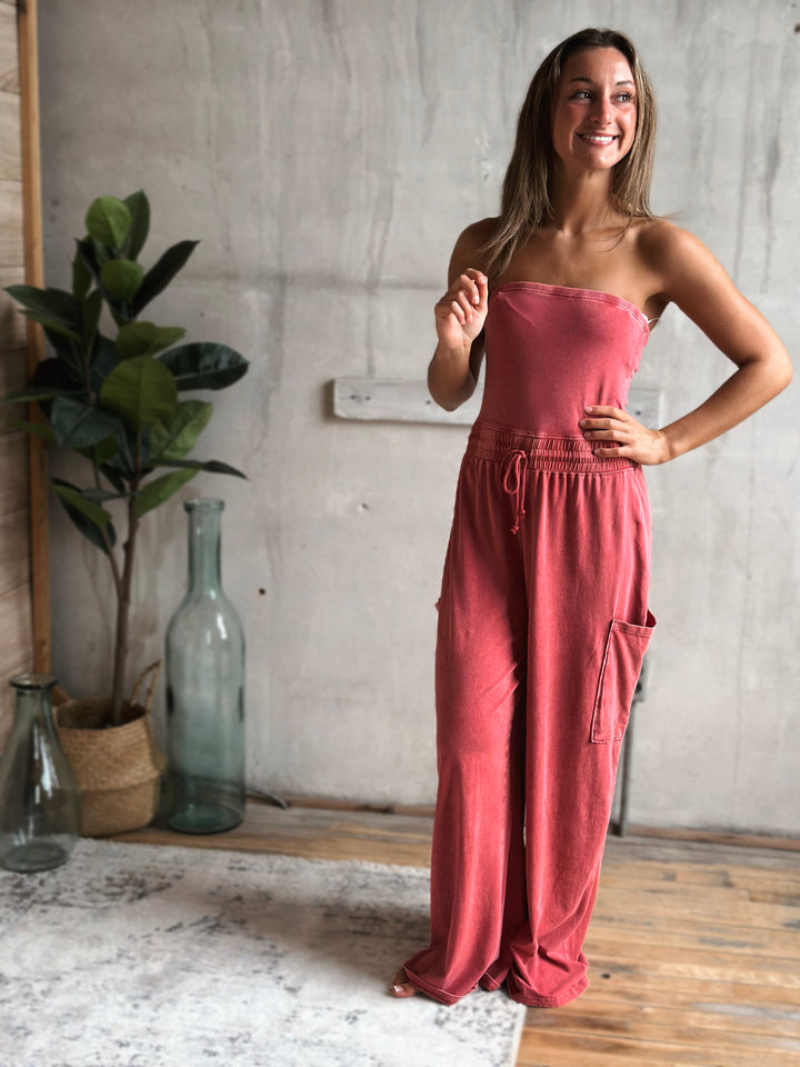 Woman wearing a light washed red strapless jumpsuit standing in a room with a plant and bottles.