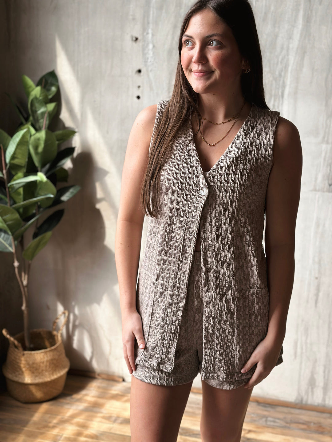 Woman wearing a textured beige sleeveless top and shorts standing indoors with a plant in the background.