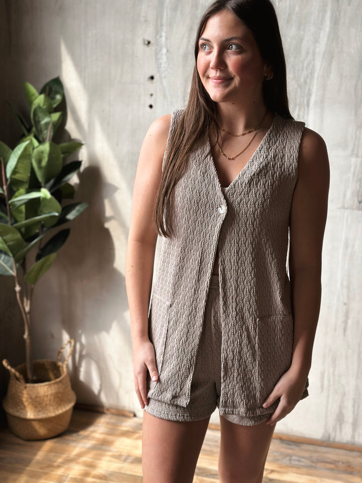 Woman wearing a textured beige sleeveless top and shorts standing indoors with a plant in the background.