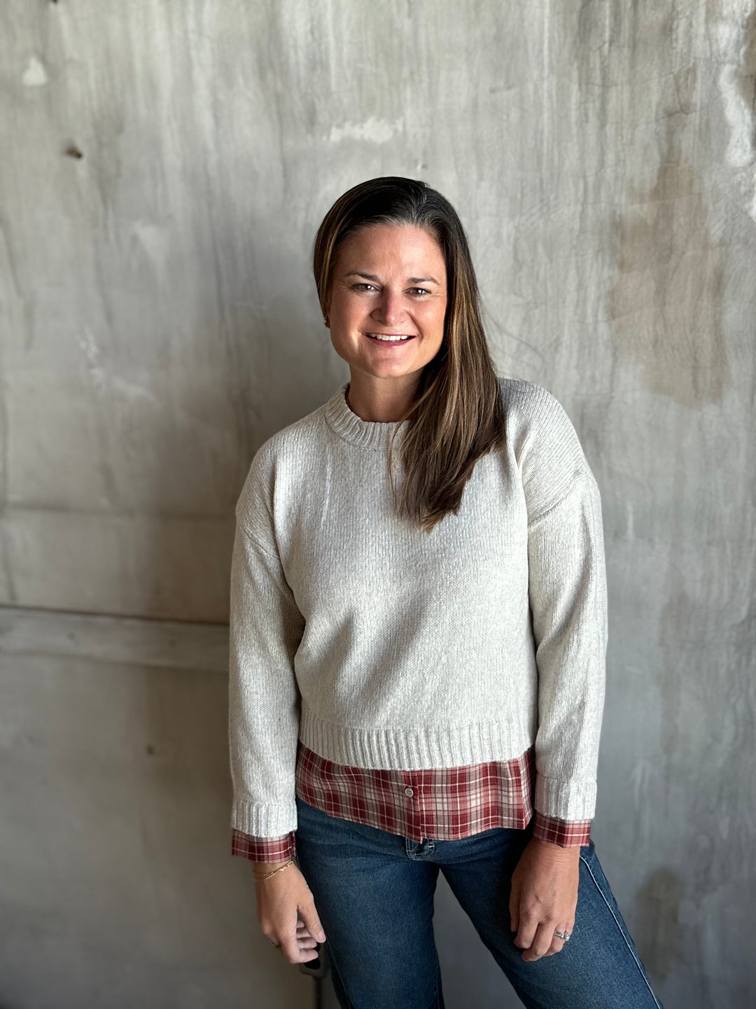 A woman wearing a cream-colored knit crewneck sweater, a red and white plaid flannel shirt, and blue jeans, standing against a textured concrete wall.
