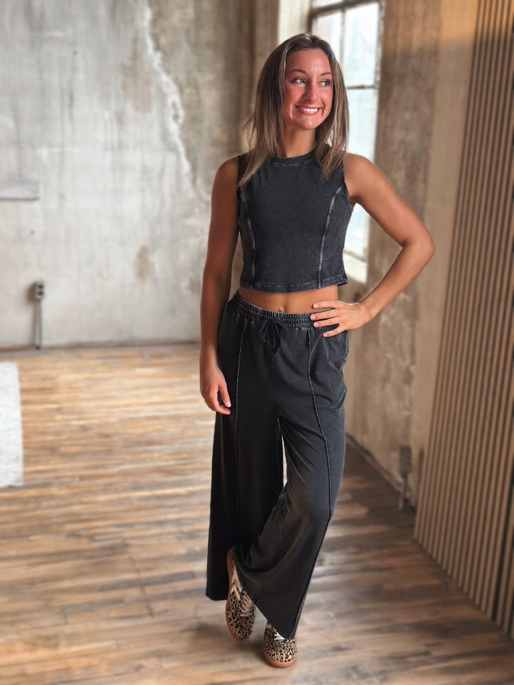 Woman wearing a black sleeveless top and black wide-leg pants in a room with wooden flooring and a textured wall.