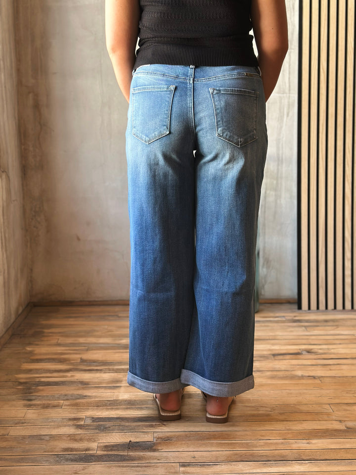 Person wearing blue jeans and a black top standing on a wooden floor with a neutral wall background