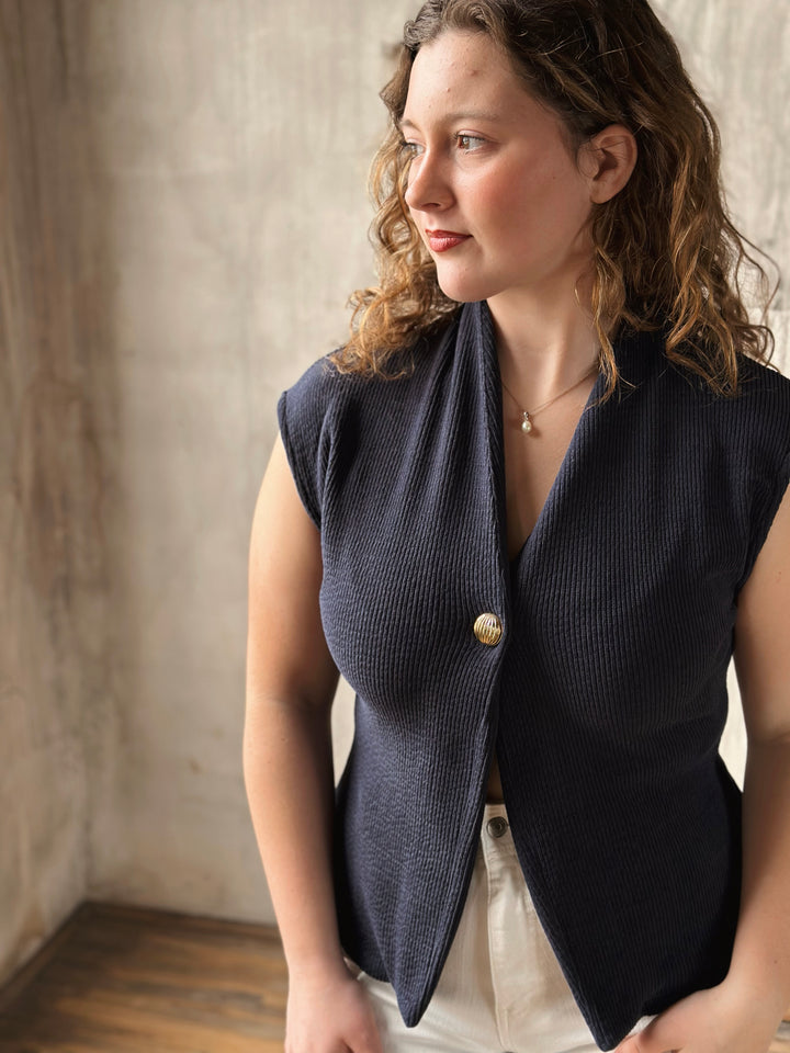 Woman wearing a navy blue sleeveless blazer with a gold button against a neutral background