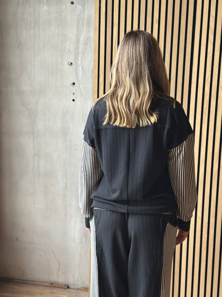 A woman viewed from behind wearing a dark charcoal ribbed jacket with striped sleeves and matching ribbed trousers, standing in front of a slatted wood wall.