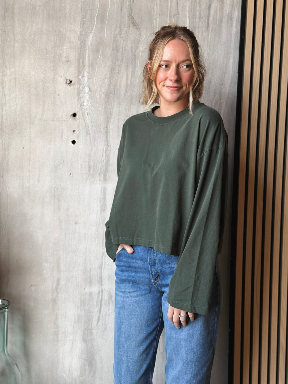 Person wearing a green sweater and blue jeans standing against a textured wall.