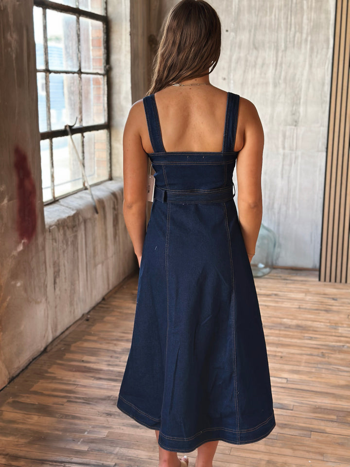 A woman wearing a dark blue denim dress standing in a room with a wooden floor and large window.