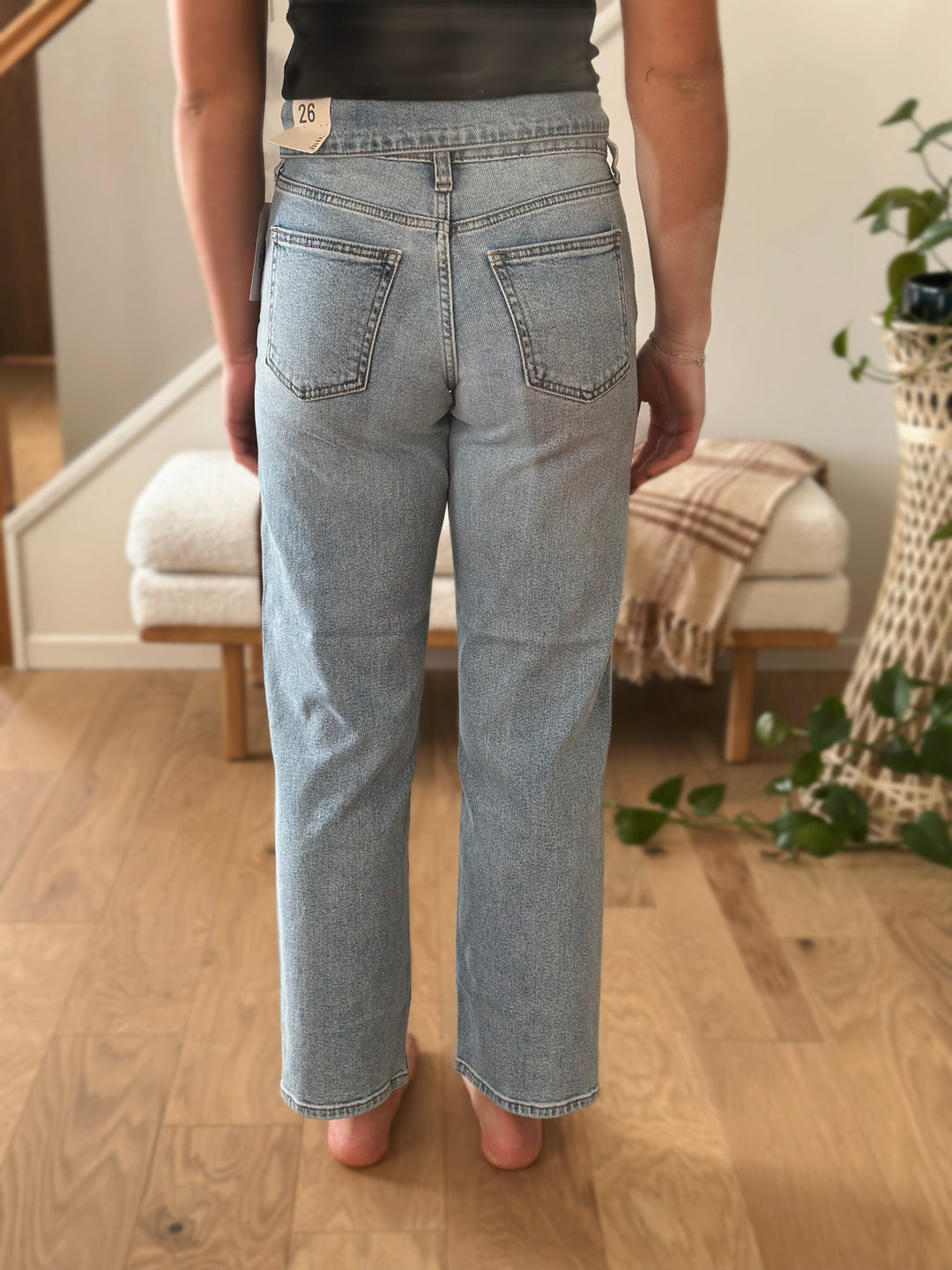 Person wearing light blue jeans standing in a room with wooden floor and plant.