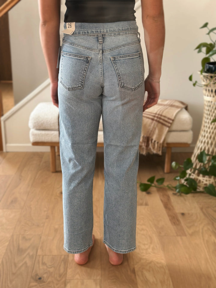 Person wearing light blue jeans standing in a room with wooden floor and plant.