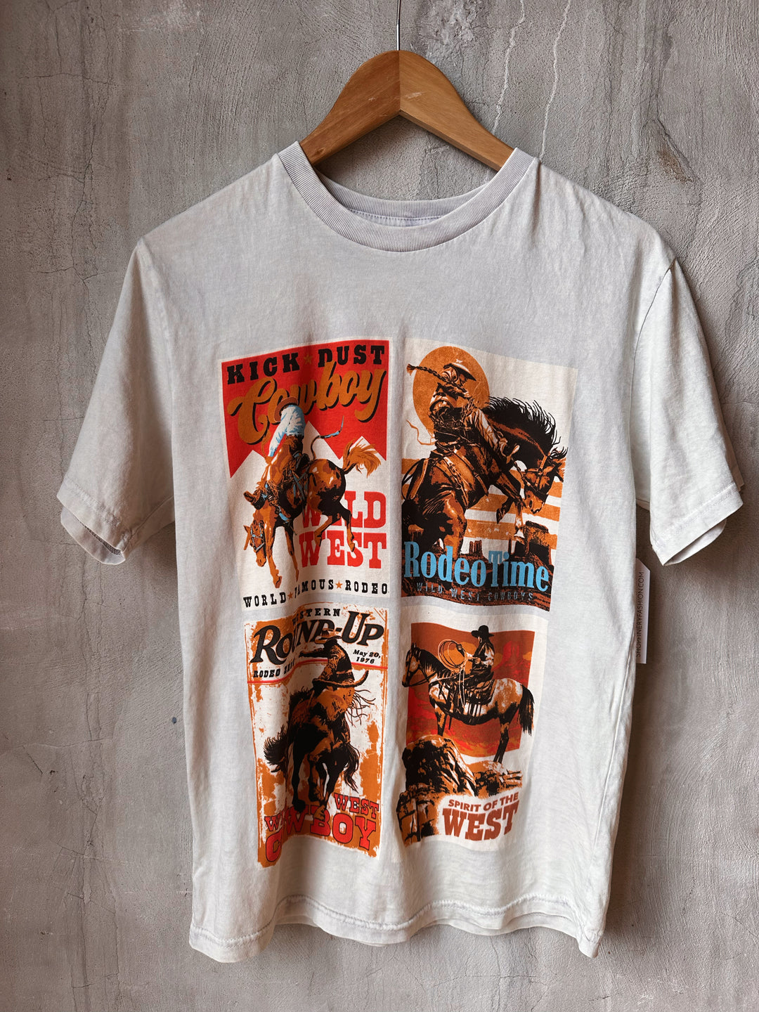White t-shirt with cowboy graphic design on a wooden hanger against a gray concrete wall.