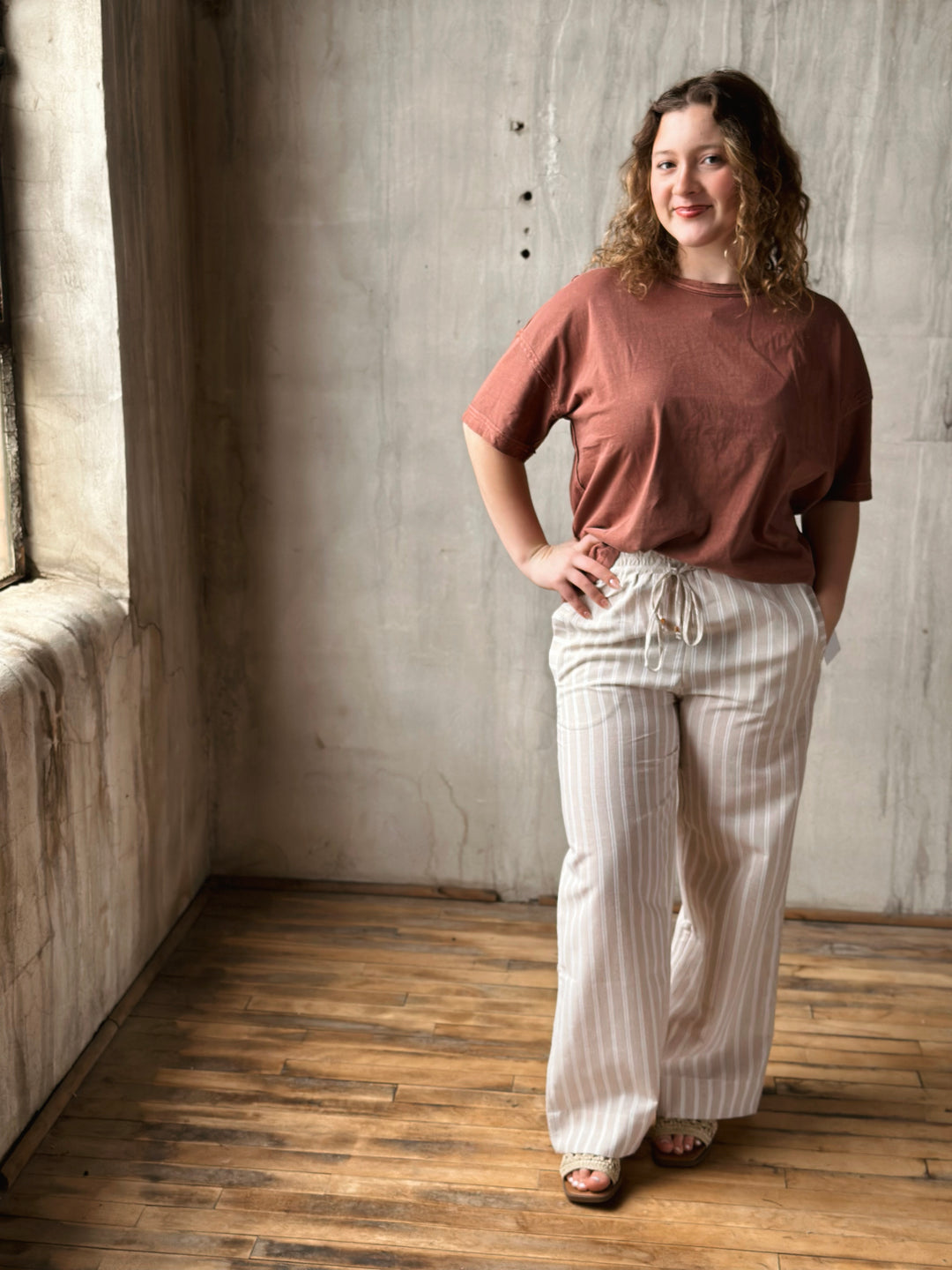 Woman wearing a brown top and white striped pants standing in a room with wooden floor and gray walls.