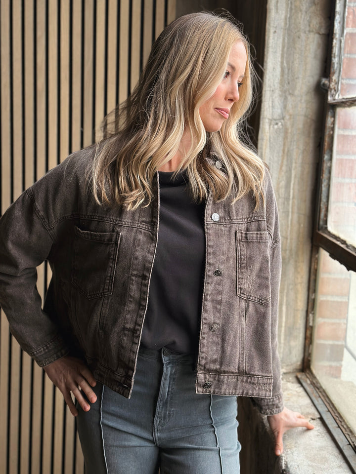 Woman wearing a black denim jacket and jeans standing in front of a window.