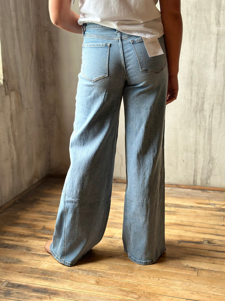 A person shown from the back wearing light-wash wide-leg denim jeans and a white top, standing barefoot on a wooden floor against a textured concrete wall.