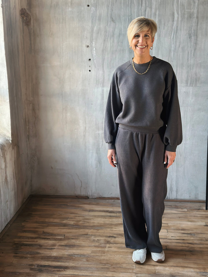 Person wearing a gray outfit standing in a room with wooden floor and concrete wall.