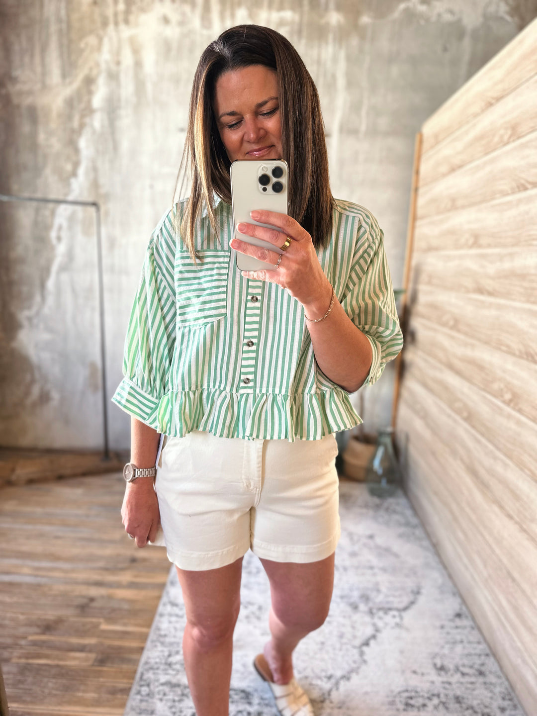 Woman taking a mirror selfie wearing a green and white striped shirt and white shorts.