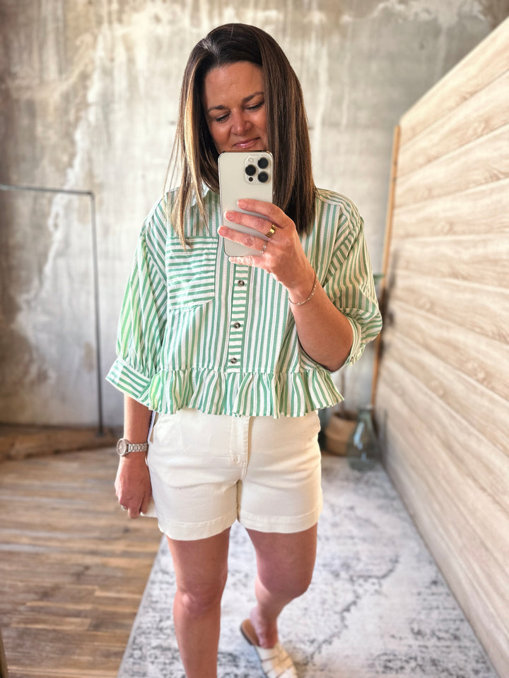 Woman taking a mirror selfie wearing a green and white striped shirt and white shorts.