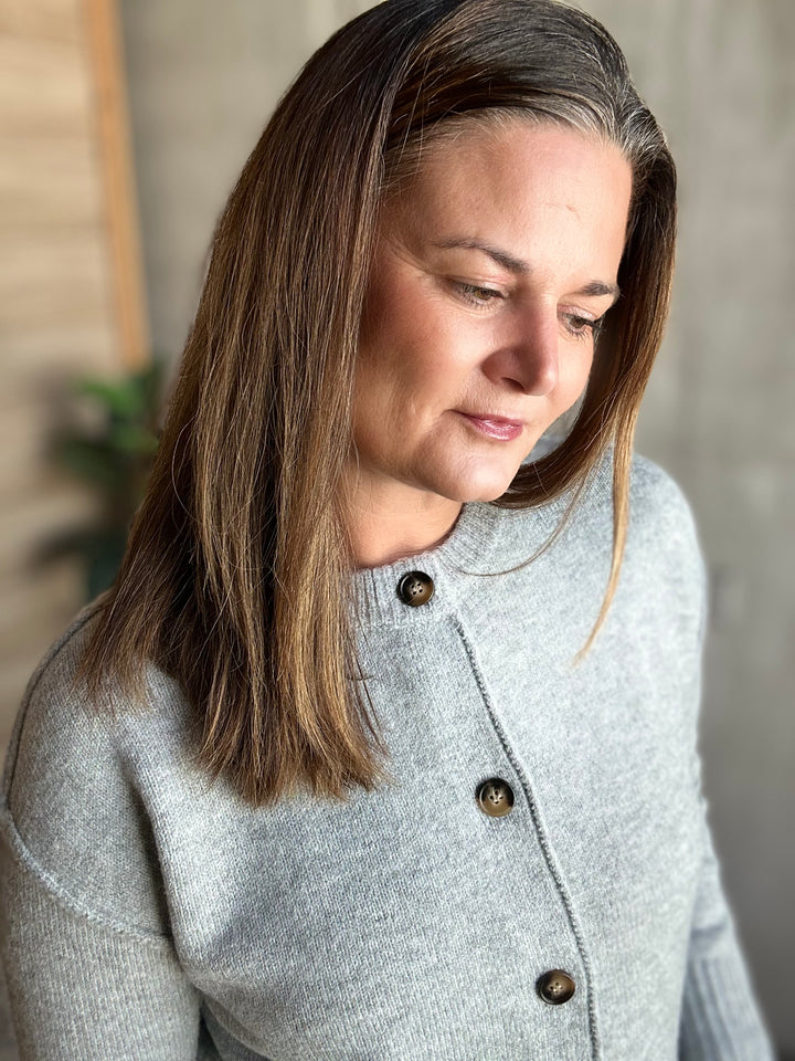 A woman with shoulder-length brown hair wearing a light gray knit cardigan with dark brown buttons, shown in a close-up portrait.